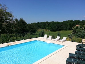 A rectangular swimming pool with clear blue water, surrounded by a tiled deck. There are several white lounge chairs aligned along the pool's edge. The pool is set against a backdrop of lush green trees and a grassy area.