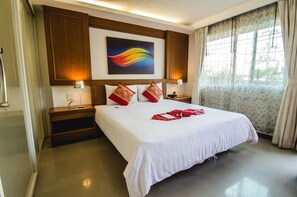 A hotel room with a large bed, wooden headboard, two bedside tables with lamps, a painting above the bed, and a window with curtains. - Patong Terrace Boutique Hotel (Patong)