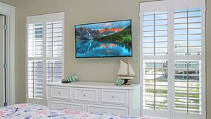 Un dormitorio con cama, televisor montado en la pared, cómoda y ventana con persianas.