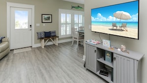 Una sala de estar con un televisor mostrando una escena de playa, un comedor con mesa y sillas, y una estantería con libros y objetos decorativos.