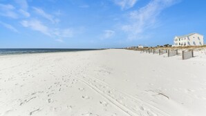 Una extensa playa arenosa con huellas que conducen a una casa blanca con balcón, con vista al océano.