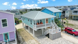 Una casa frente a la playa con un porche cubierto, una escalera que lleva a una terraza y una camioneta roja estacionada al frente.
