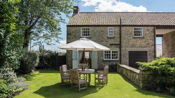 A stone building with a tiled roof, a patio with a table and chairs, and a well-maintained lawn.