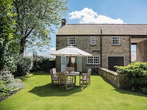 A stone building with a tiled roof, a patio with a table and chairs, and a well-maintained lawn.
