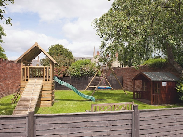 A wooden play structure with a slide, a small wooden house, and a brick wall.