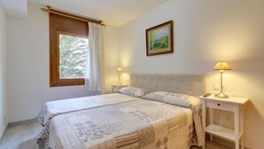 Quarto com uma cama grande, duas mesas de cabeceira, um criado-mudo, uma janela com cortinas, um quadro na parede e uma luminária no criado-mudo.
