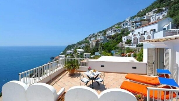 Eine Terrasse mit Tisch und Stühlen, einem Blumentopf und Blick auf das Meer sowie Hügelhäuser.