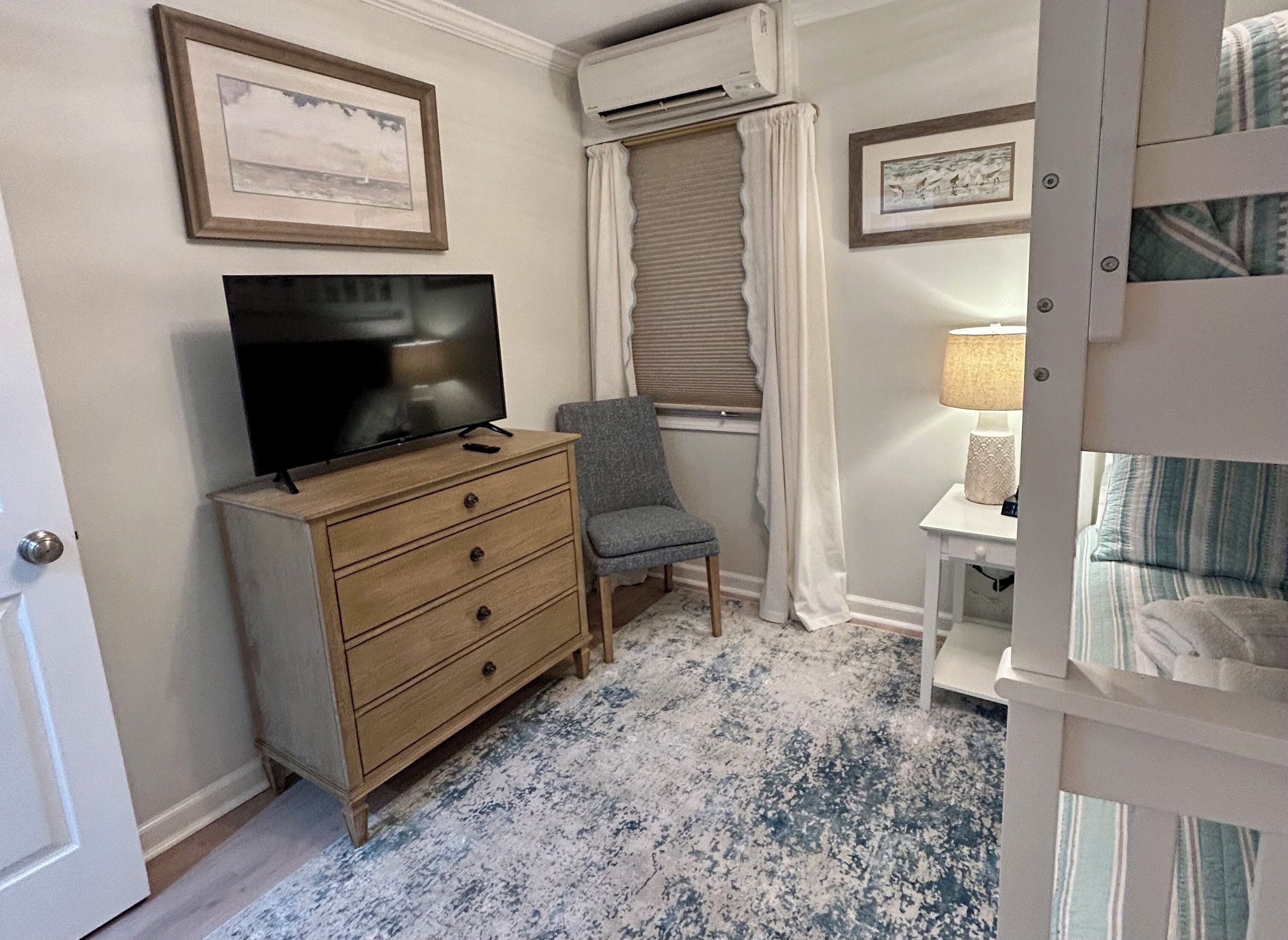 A bedroom with a television on a dresser, a chair, a window with curtains, and a bedside table with a lamp.