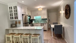 A modern kitchen with white cabinets, a stainless steel refrigerator, and a tiled backsplash. There are four bar stools at the island and a dining table with chairs.