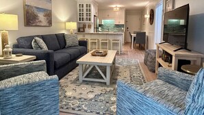 A living room with a sofa, coffee table, and patterned rug. There is a kitchen area with cabinets and a dining table. A ceiling fan is visible, and a television is mounted on the wall.