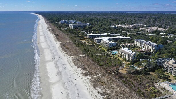 An aerial view of a coastal area with a beach, several multi-story buildings, and a swimming pool.
