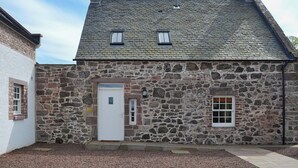 A stone building with a white door, two windows, and a chimney. - Grieve's Cottage - UK11107 (Montrose)