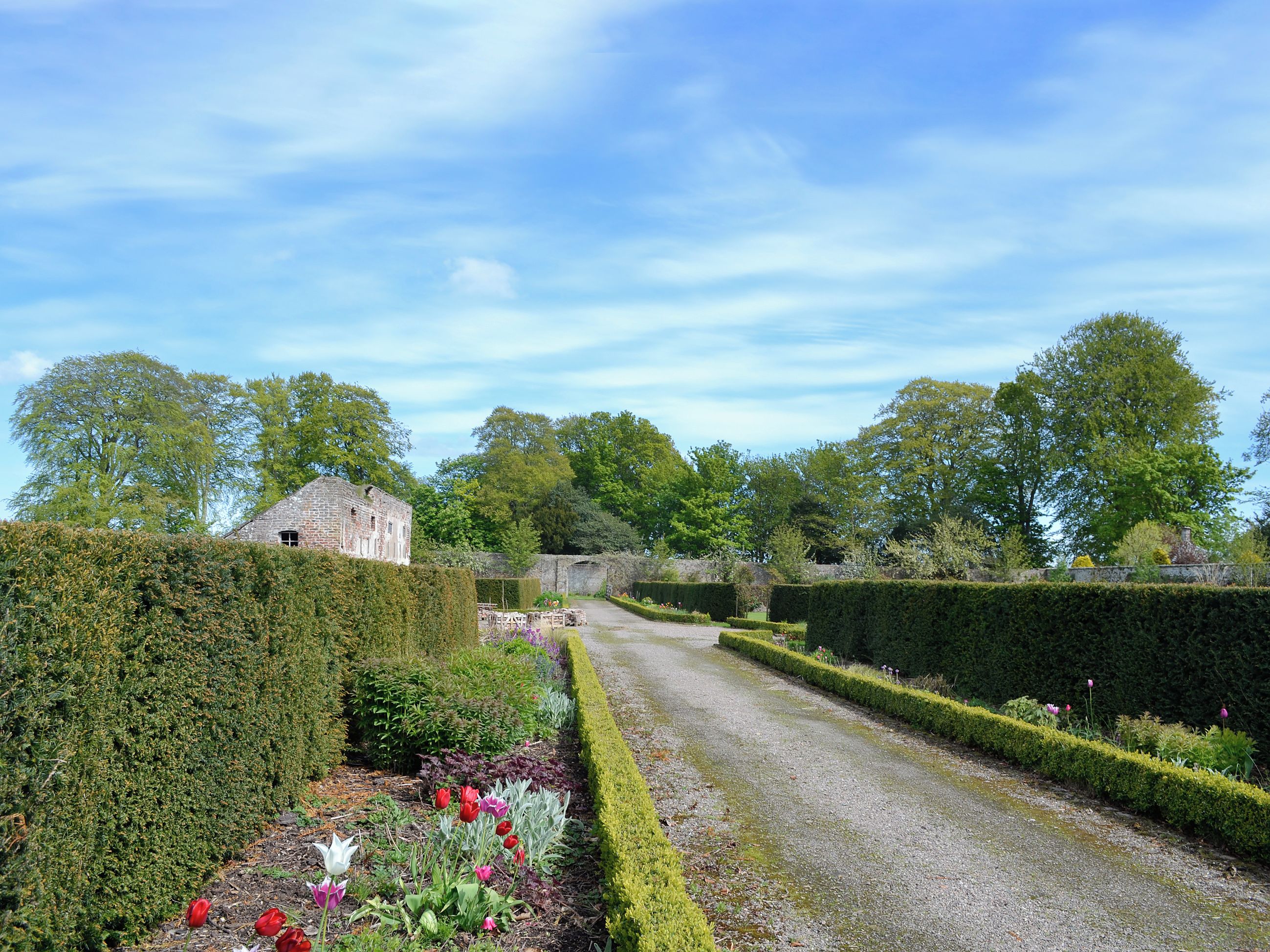 A garden pathway leading to a stone building, surrounded by neatly trimmed hedges and various plants.