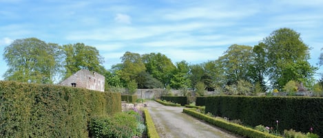 A garden pathway leading to a stone building, surrounded by neatly trimmed hedges and various plants.