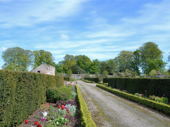 A garden pathway leading to a stone building, surrounded by neatly trimmed hedges and various plants.