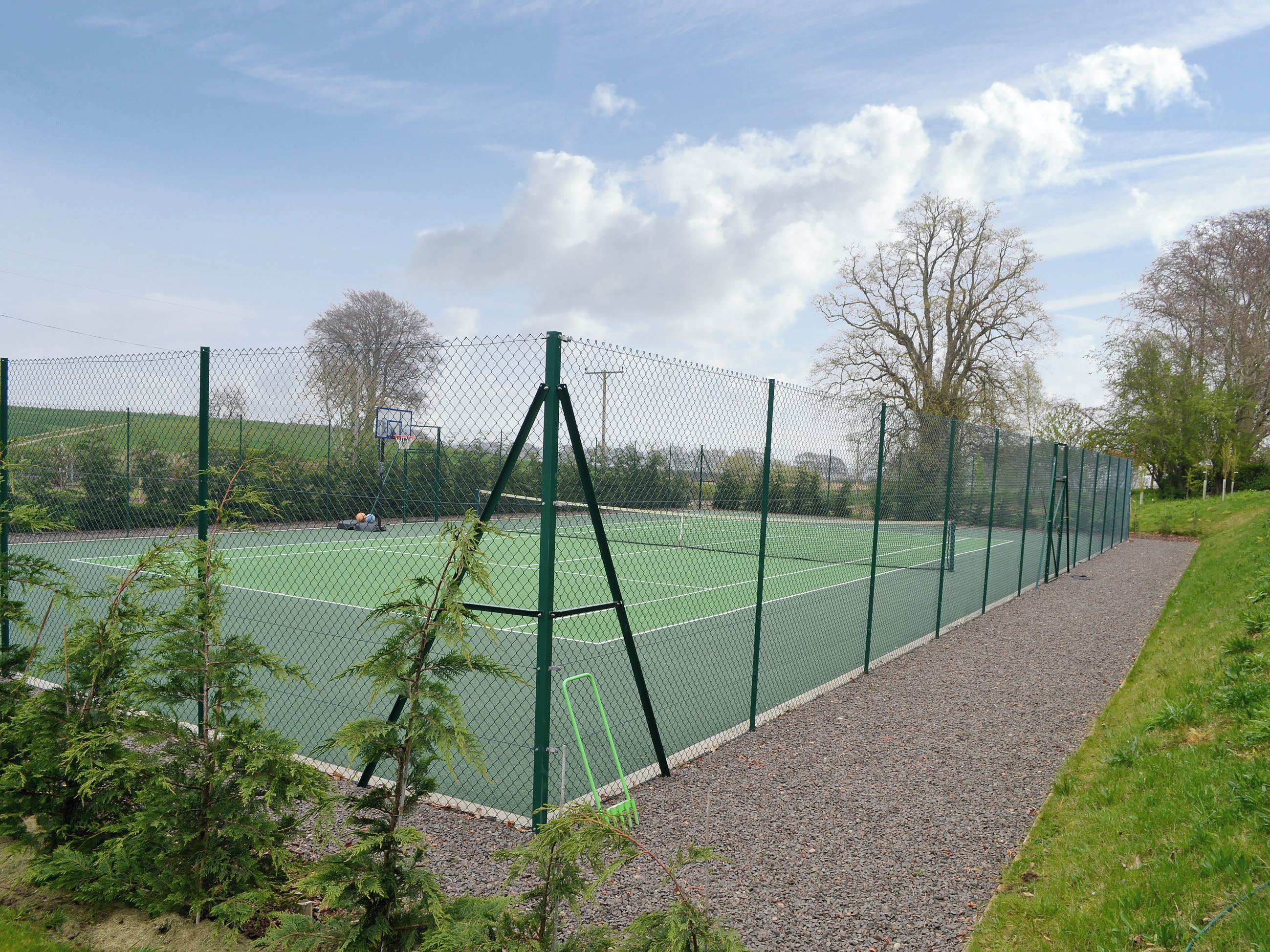 Ein Tennisplatz mit grünem Zaun, Bäumen und blauem Himmel.