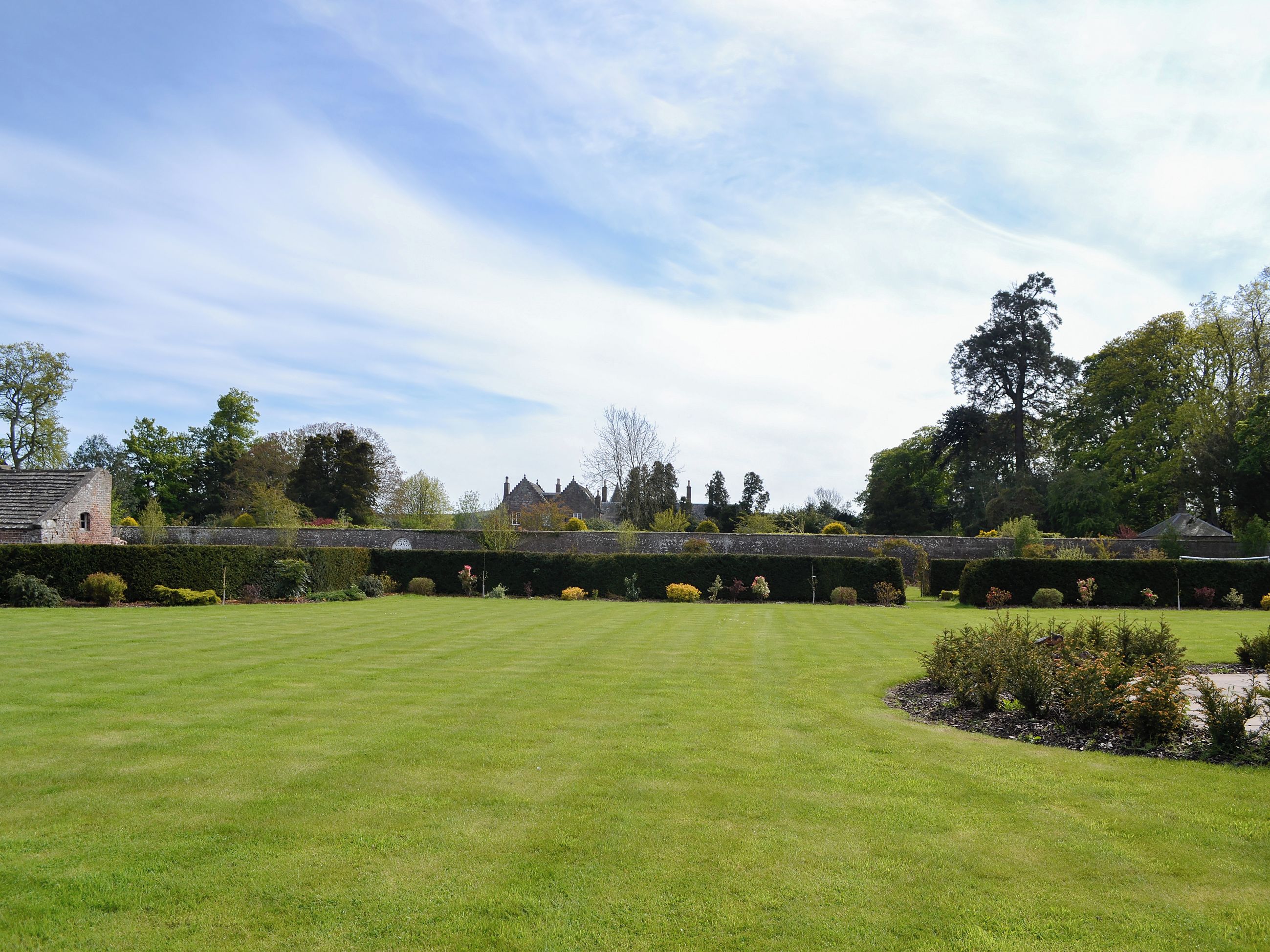 A well-maintained lawn with a stone wall and a hedge in the foreground, and a building with a traditional design in the background.