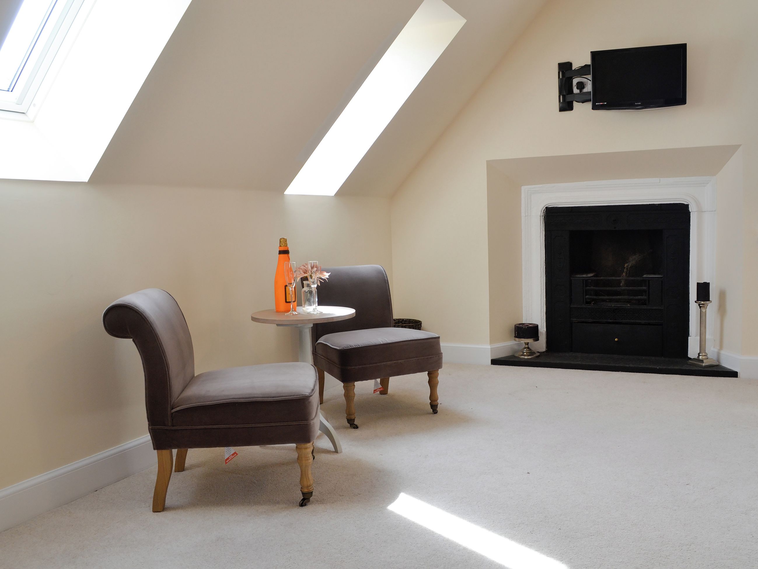 Une pièce avec un plafond haut, une cheminée, deux fauteuils, une petite table avec une bouteille et des verres, et un téléviseur fixé au mur.