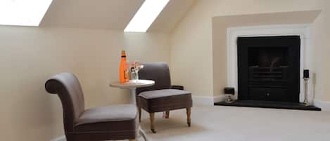 Une pièce avec un plafond haut, une cheminée, deux fauteuils, une petite table avec une bouteille et des verres, et un téléviseur fixé au mur.