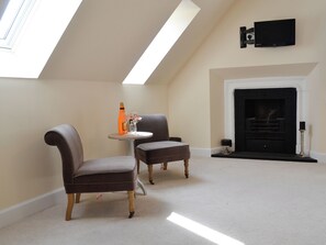 Een kamer met een hoge plafond, een open haard, twee fauteuils, een kleine tafel met een fles en glazen, en een bevestigde televisie.