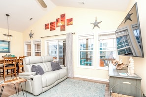 A living room with a sofa, a dining table, and chairs, a large window with curtains, and wall art.