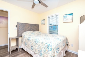 Quarto com cama, ventilador de teto, janela com persianas e uma mesa pequena.