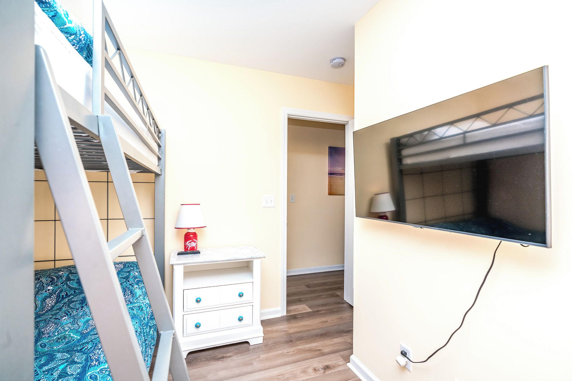 A room with a bunk bed, a small white nightstand with a lamp, a television mounted on the wall, and a wooden floor.