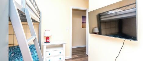 A room with a bunk bed, a small white nightstand with a lamp, a television mounted on the wall, and a wooden floor.