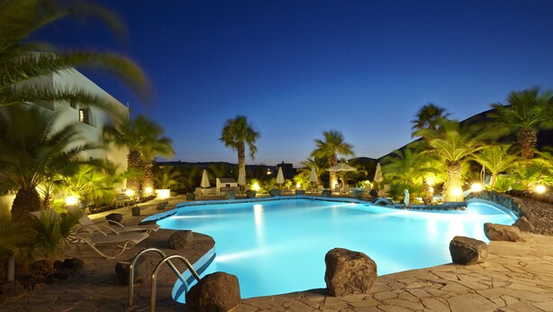 A swimming pool with lounge chairs and umbrellas, surrounded by palm trees and rocks. - Crystal Palace (Santorini)