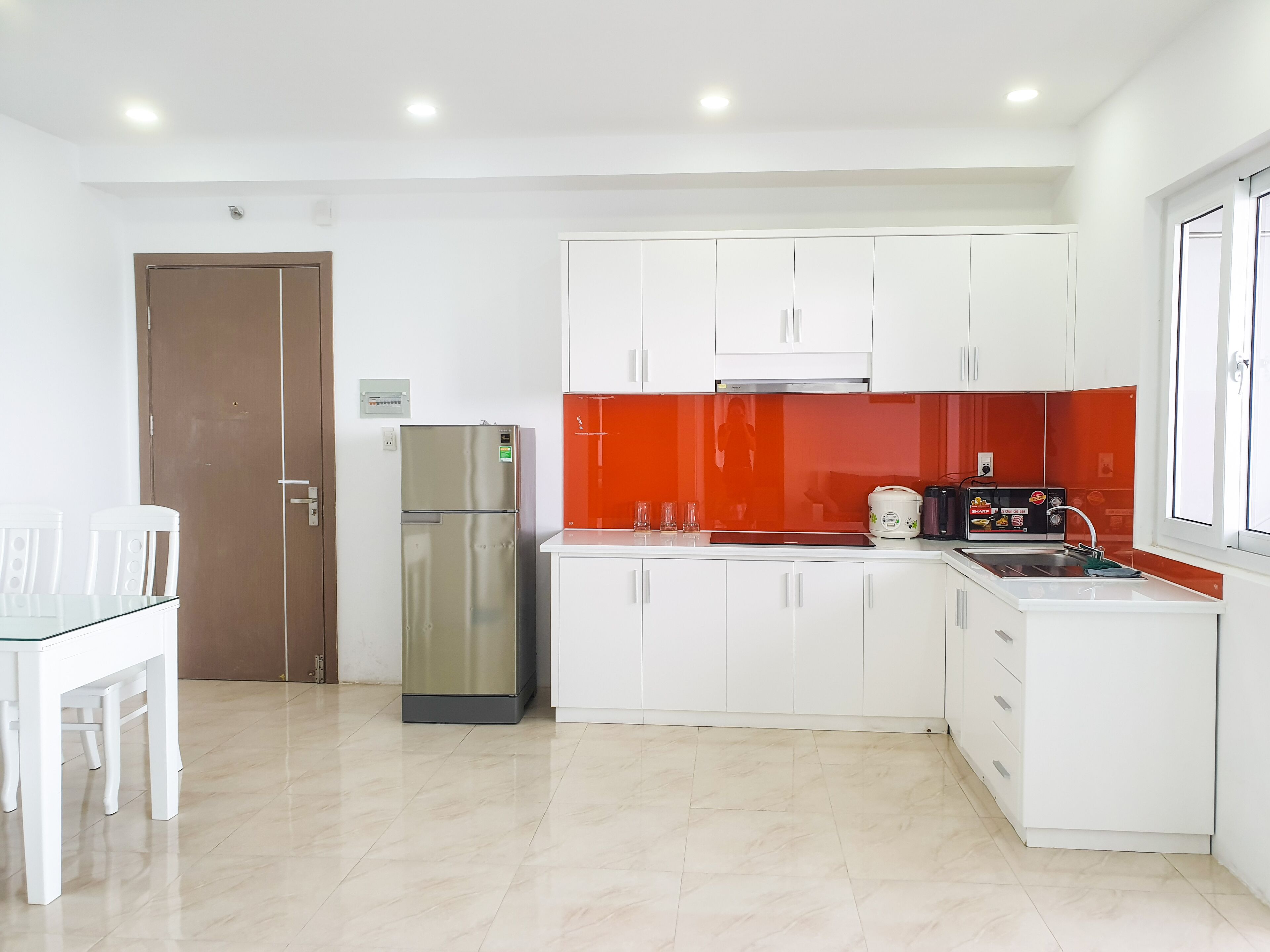Uma cozinha moderna com armários brancos, uma geladeira de aço inoxidável, um painel de parede vermelho e uma pequena área de jantar com mesa e cadeiras de vidro.