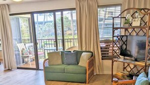 A spacious living area with a green sofa, wooden flooring, a ceiling fan, and a TV on a wooden stand. - Kauai Beach Villas D-9 ... Centrally located ... & Lagoon view (Lihue)
