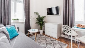 A modern bedroom with a grey sofa, a small white table, a potted plant, a white dresser, and a wall-mounted TV. - Apartamenty Pod Wawelem Wroblewskiego (Kraków)