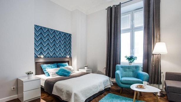 A modern bedroom with a bed, a nightstand, a blue armchair, a small table with a cup, and a window with curtains. - Apartamenty Pod Wawelem Wroblewskiego (Kraków)