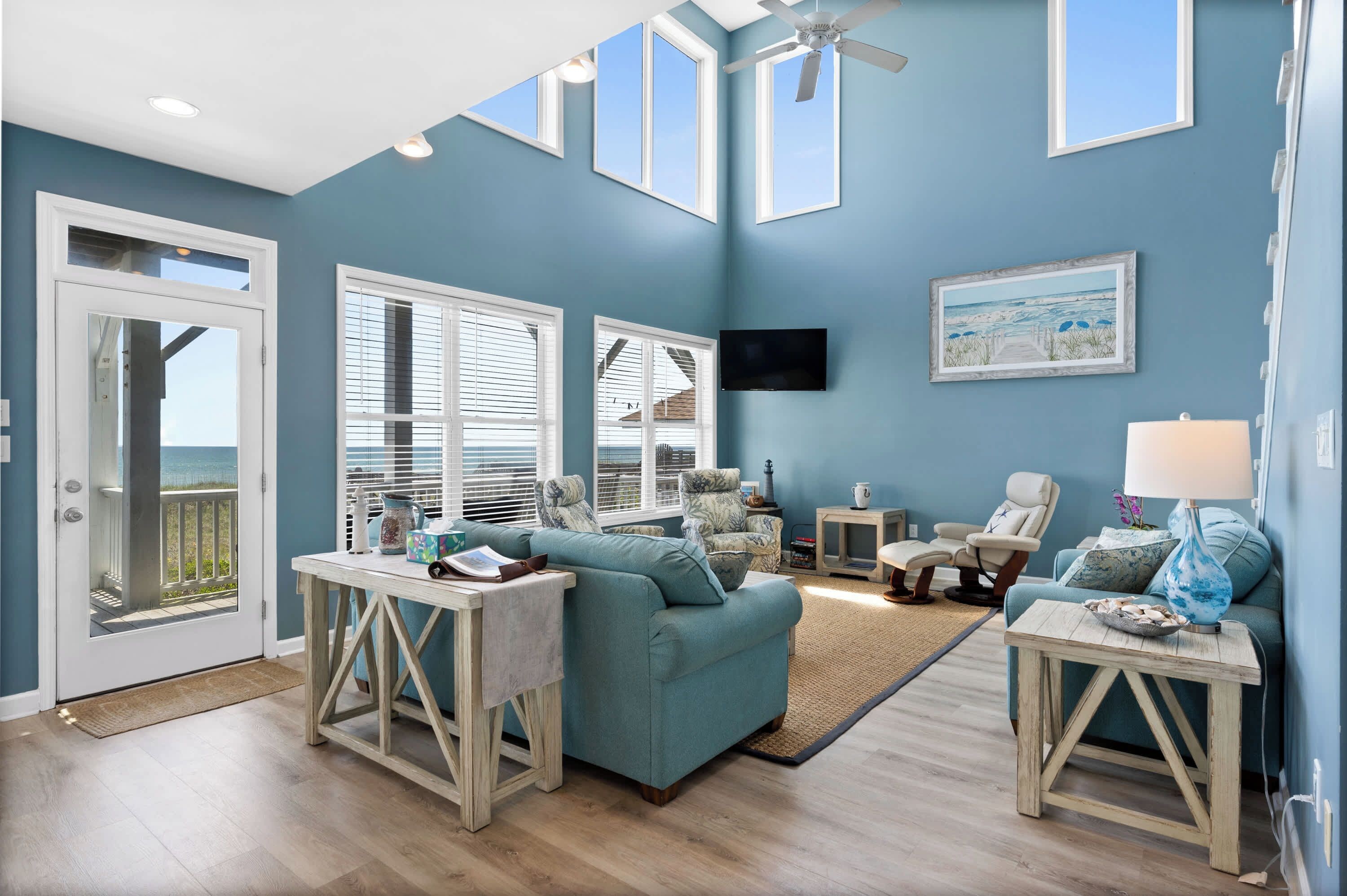 A living room with a teal sofa, a TV, a ceiling fan, and a view of the ocean through large windows.
