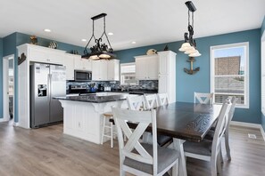 A spacious kitchen with white cabinetry, a central island, and a dining area with a dark wood table and white chairs. - Pirate`s Booty by Brunswick Vacation Rentals: OIB 5BR Oceanfront Home w/ Pool (Ocean Isle Beach)