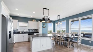 A kitchen with white cabinets, a central island, a dining area with chairs, and pendant lights. - Pirate`s Booty by Brunswick Vacation Rentals: OIB 5BR Oceanfront Home w/ Pool (Ocean Isle Beach)