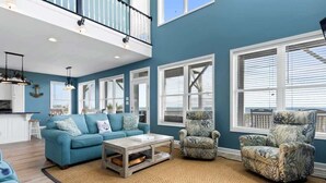 A spacious living room with a high ceiling, large windows, a blue sofa, a patterned armchair, and a coffee table. - Pirate`s Booty by Brunswick Vacation Rentals: OIB 5BR Oceanfront Home w/ Pool (Ocean Isle Beach)