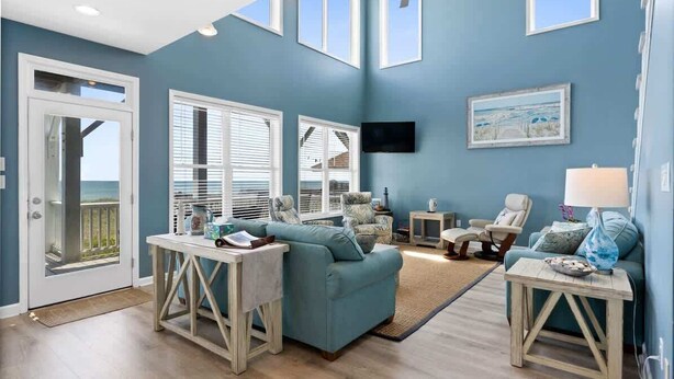 A living room with a blue wall, a ceiling fan, a TV mounted on the wall, and a view through the glass door. - Pirate`s Booty by Brunswick Vacation Rentals: OIB 5BR Oceanfront Home w/ Pool (Ocean Isle Beach)