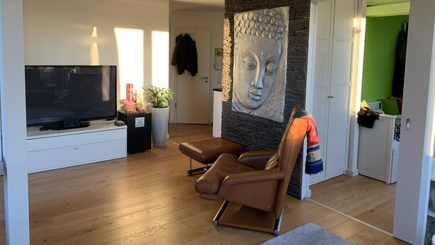 Een moderne woonkamer met een flatscreen televisie, leren fauteuil, een potplant en een groot wandsculptuur.