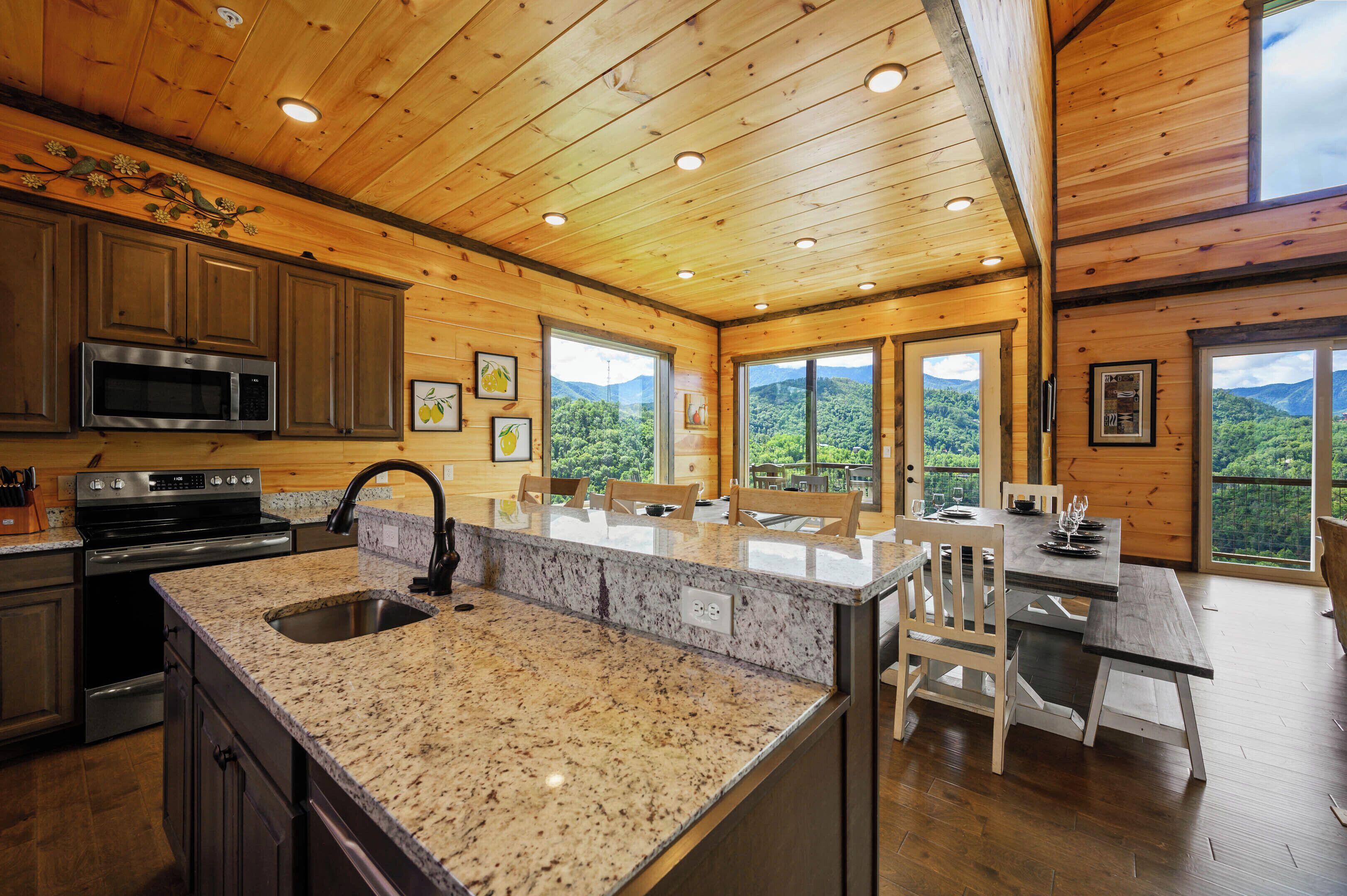 A kitchen with granite countertops, stainless steel appliances, and wooden cabinets. There is a large island with seating, and the room has large windows offering a view of the mountains.
