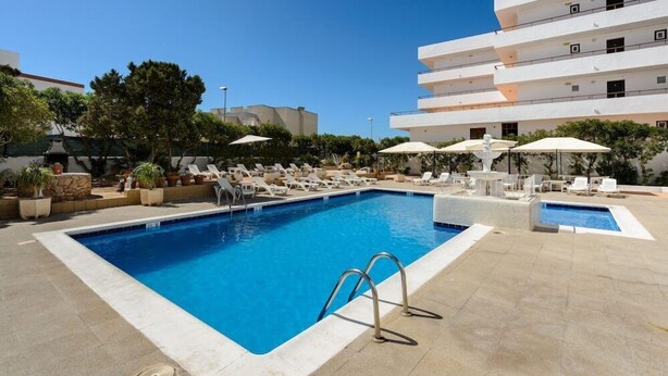A swimming pool with lounge chairs and umbrellas, surrounded by a building with multiple balconies. - Apartamentos San Antonio Beach (Sant Josep de sa Talaia)