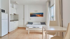 A compact living space with a kitchenette, a sofa with patterned cushions, a dining table, and a painting on the wall. - Apartamentos San Antonio Beach (Sant Josep de sa Talaia)