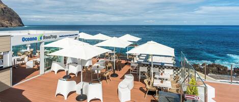 A rooftop bar with white umbrellas, outdoor seating, and a view of the ocean.