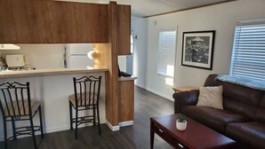 Een moderne woonkamer met een bruine leren bank, een houten salontafel en twee metalen stoelen. In de achtergrond is een keuken te zien met een houten kast en een aanrecht.