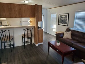 A modern living room with a brown leather sofa, a wooden coffee table, and two metal chairs. A kitchen area with a wooden cabinet and a countertop is visible in the background. - Charming Riverfront Escape with Private Dock on the Colorado (Mohave Valley)