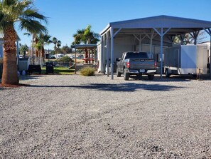 A gravel parking area with a covered carport, a truck, and a building with a metal roof. - Charming Riverfront Escape with Private Dock on the Colorado (Mohave Valley)