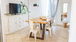 Un comedor con una mesa de madera y sillas blancas, un televisor en un mueble, y una planta.