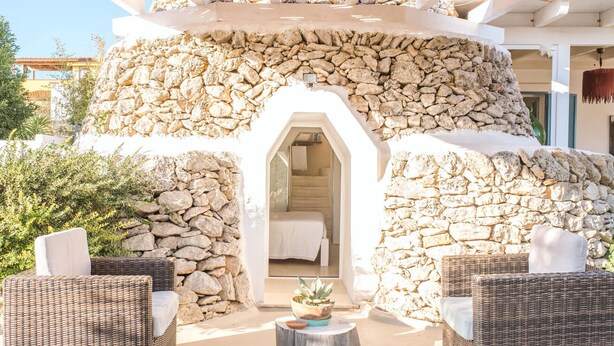 Una estructura de paredes de piedra con un acceso a un dormitorio, muebles de mimbre y una pequeña mesa con una planta.
