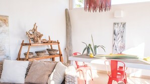 Un salón luminoso con un sofá blanco, sillas rojas, un estante de madera con cestas y una mesa de comedor con un florero y plantas.