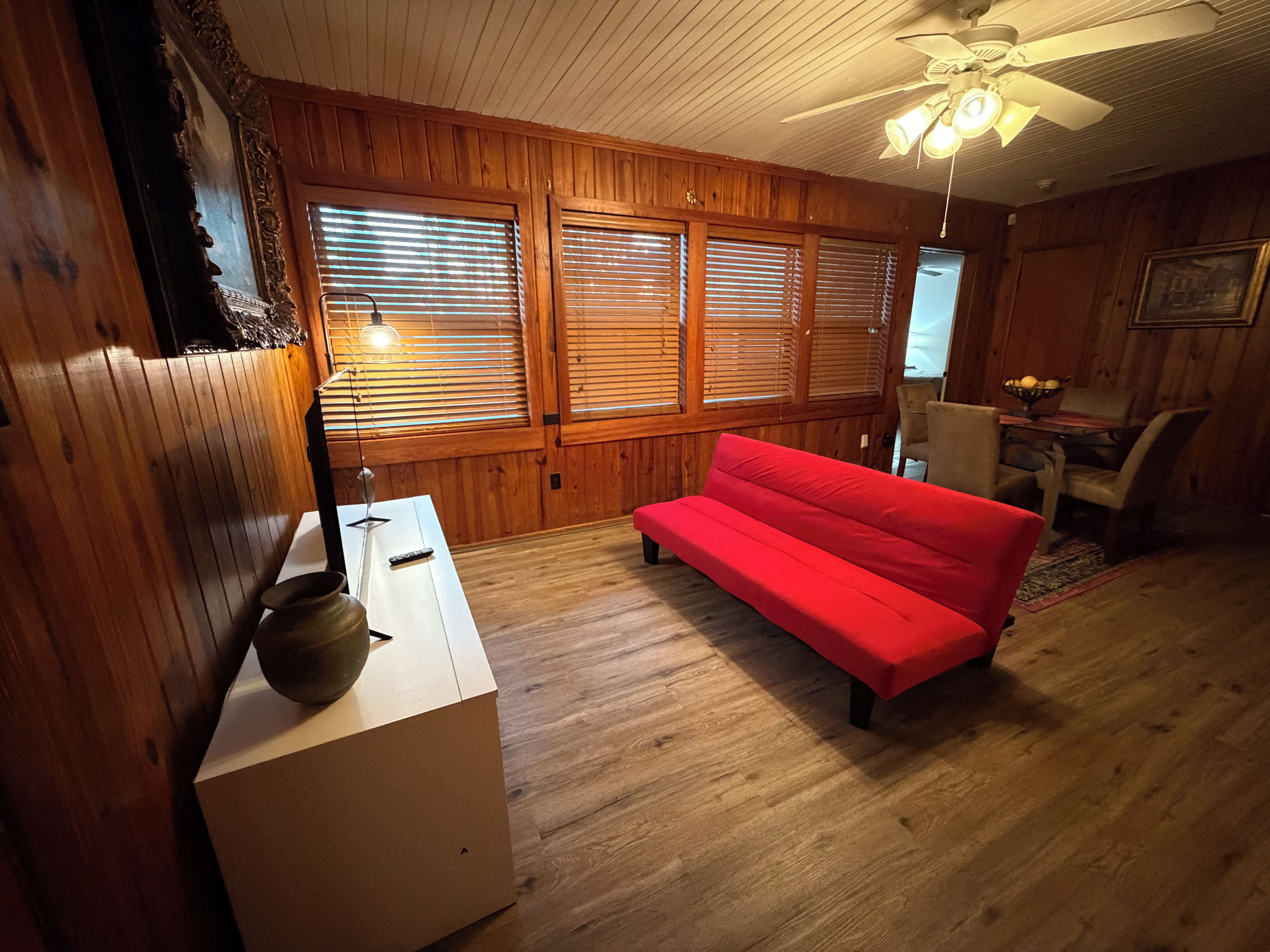 Une pièce avec un plancher en bois, un canapé rouge, une table blanche ornée d’un vase et un ventilateur de plafond.